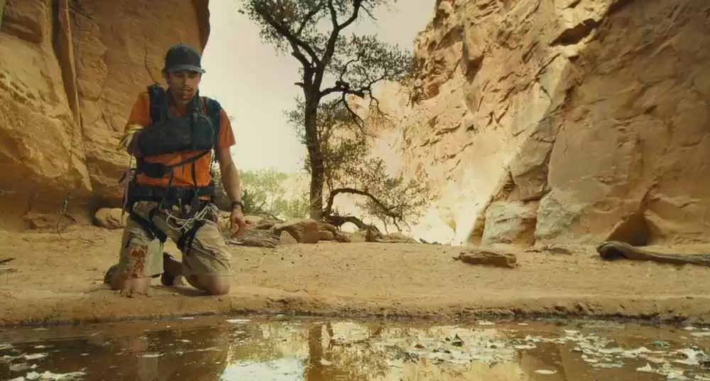 A bloodied, exhausted-looking James Franco, one arm in a sling, kneels by a leaf-covered pond in a rocky valley in 127 Hours.